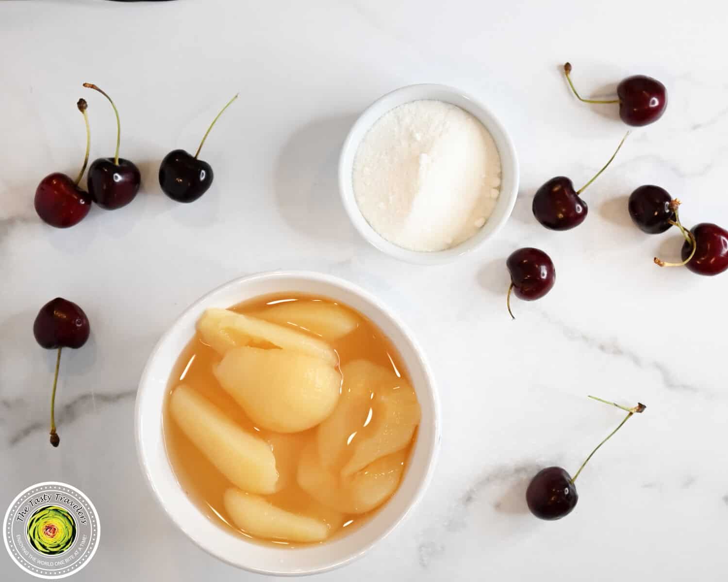 Fresh cherries with pear halves in syrup and a bowl of sugar on a white marble surface.