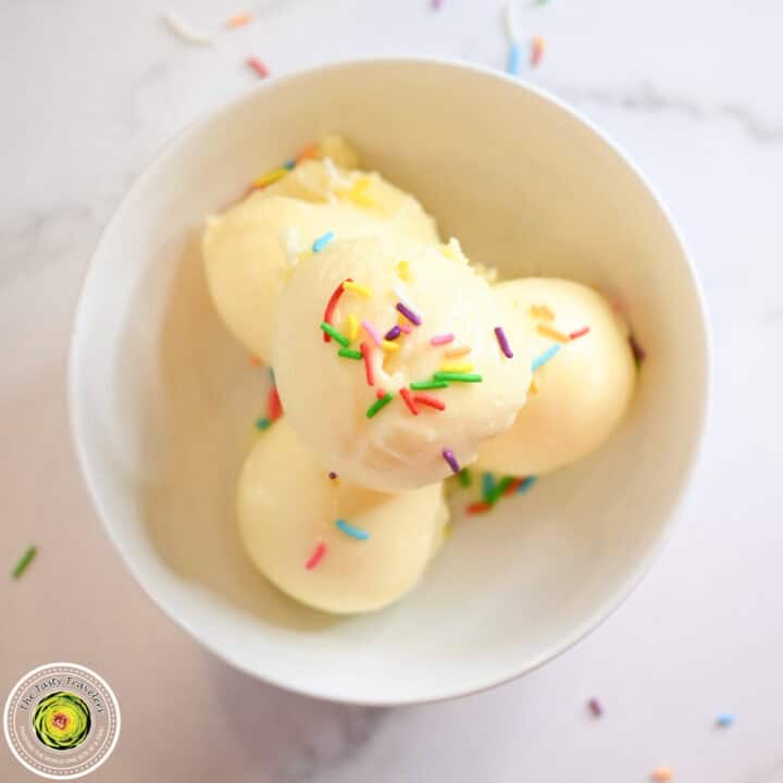 Ice cream balls with colorful sprinkles in a white cup on a white surface.
